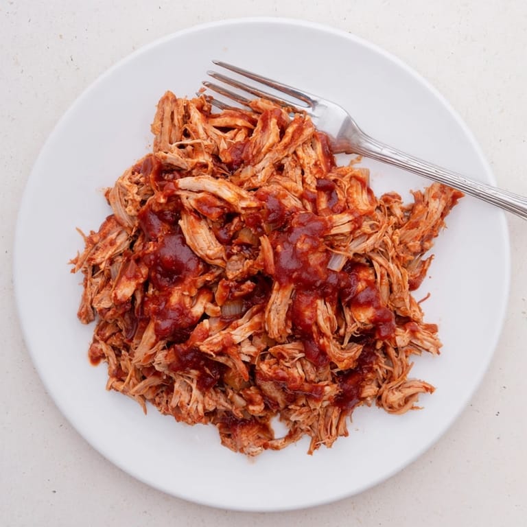 A close-up of saucy BBQ Pulled Chicken in a bowl, garnished with fresh parsley and pickles.